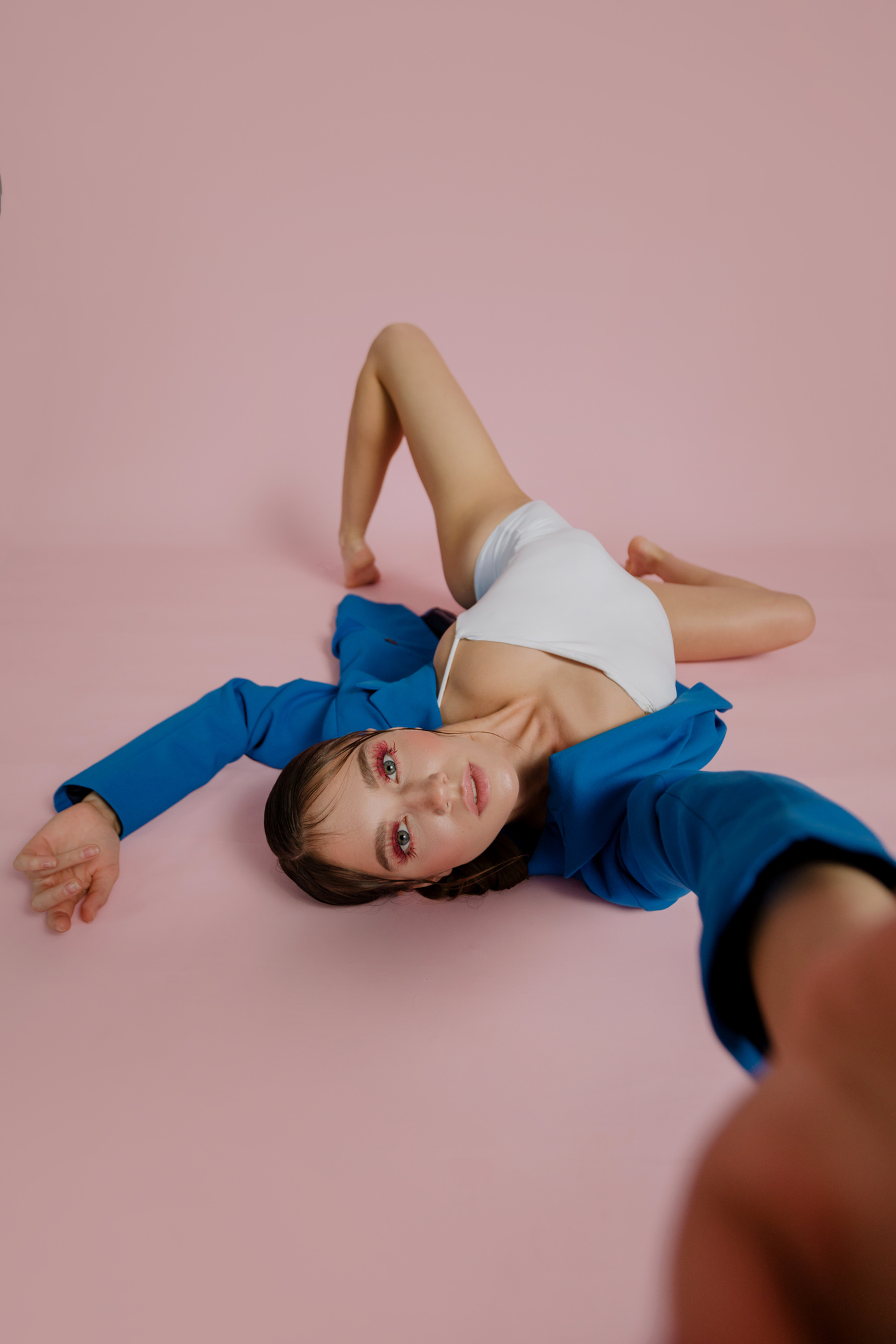 A Woman in White Spaghetti Strap and Blue Coat Lying on a Pink Surface while Looking at the Camera
